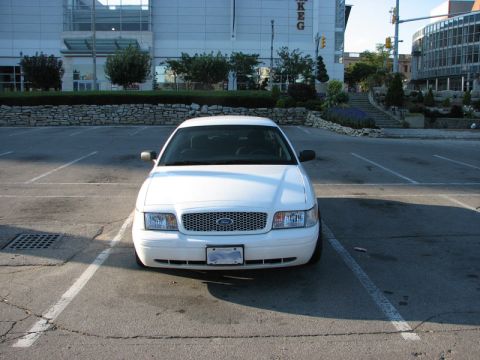 2003 Ford Crown Victoria - Head-on front view of a white Ford Crown Victoria sedan parked in an empty city lot, showing a mesh grille with Ford oval, clear headlights…
