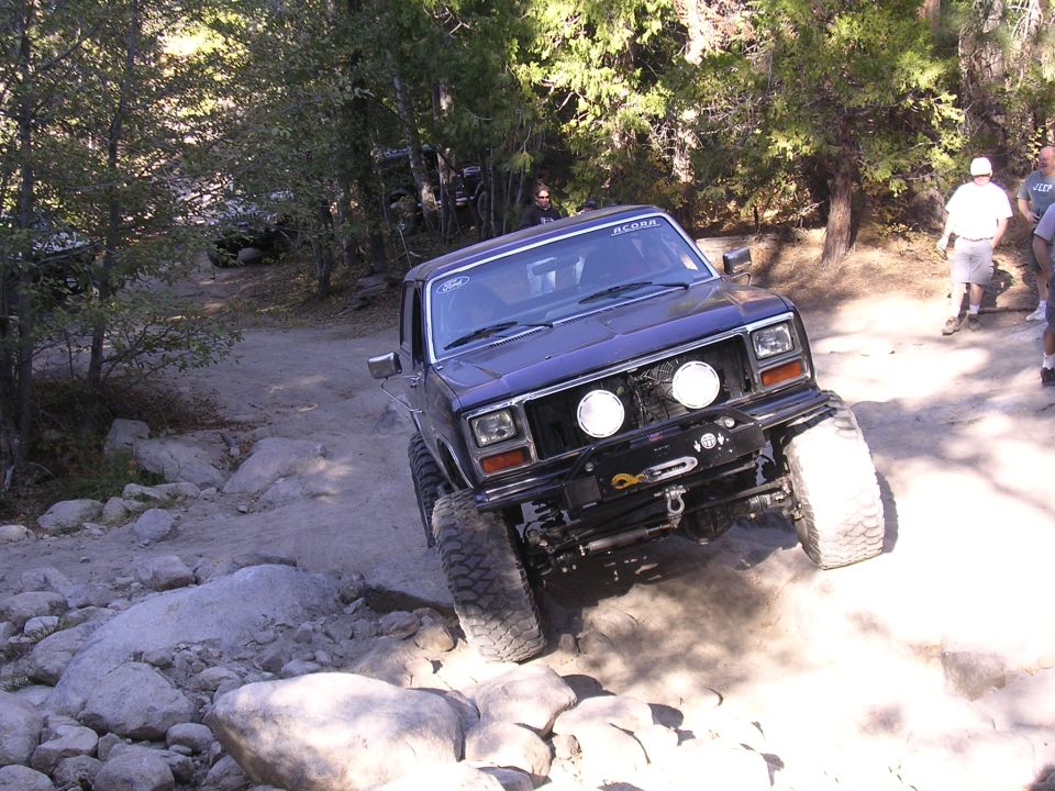 Dark blue off-road truck with large tires climbing over rocks on a dirt trail. Has two round headlights and a winch on the front