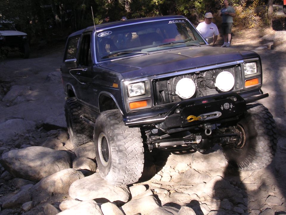 Dark blue Ford Bronco with large tires on rocks, front view. Winch and auxiliary lights visible