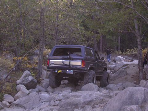 1985 Toyota 4Runner - This SUV is shown from the rear, tackling a challenging rocky trail in a wooded area.