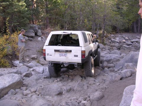 1985 Toyota 4Runner - A white 1985 Toyota 4Runner is shown from the rear, tackling a challenging rocky terrain.