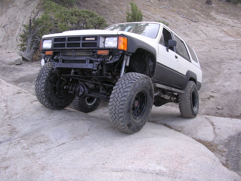 White Toyota 4Runner with huge tires climbing rocks. It has a lifted suspension and aftermarket bumper
