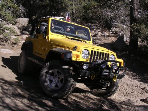 1985 Toyota 4Runner - Yellow Jeep Wrangler Rubicon navigates an off-road trail with trees and rocks in the background.