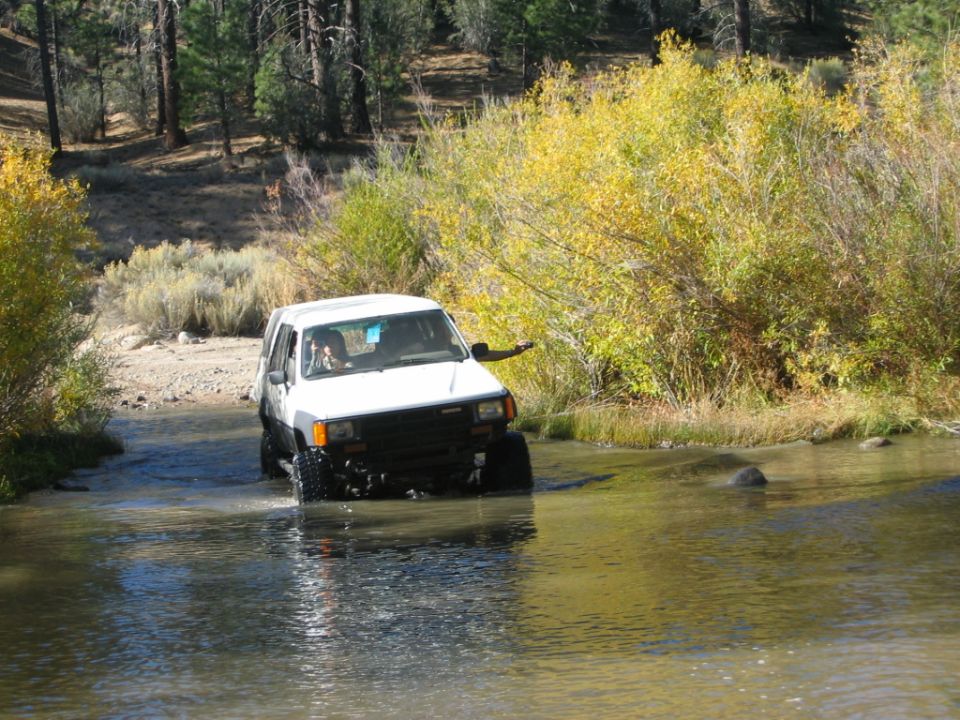 White lifted SUV crossing a shallow river with autumn brush behind