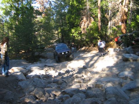 1985 Ford Bronco - Several off-road vehicles traverse a challenging, rocky path through a wooded area.