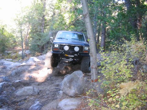 1985 Ford Bronco - Off-road SUV navigating a challenging rocky trail in a wooded area.
