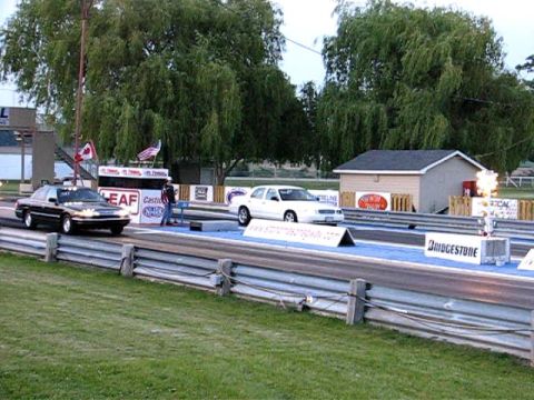 2003 Ford Crown Victoria - Two sedans are lined up on a drag strip, ready for a race, with a small building and trees visible.