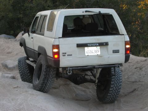1985 Ford Bronco - This white Toyota 4Runner is equipped with large off-road tires and is seen from the rear on a sandy terrain.