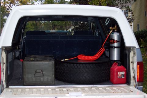 1987 Ford Bronco - The cargo area of a white SUV contains a spare tire, a metal toolbox, a red gas can, and a CO2 tank with a red hose.