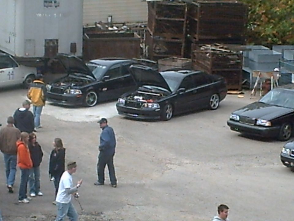 Several people stand around three black cars, two with hoods open, in an outdoor lot with industrial bins