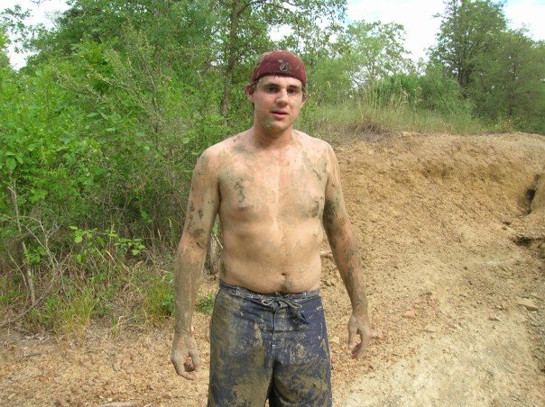 Mud-covered man in a red cap and blue shorts stands in a dirt area with trees behind him