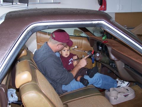 1973 Ford Ranchero - A man holds a baby while sitting in the driver's seat of a 1973 Ford Ranchero, pointing at the steering column.
