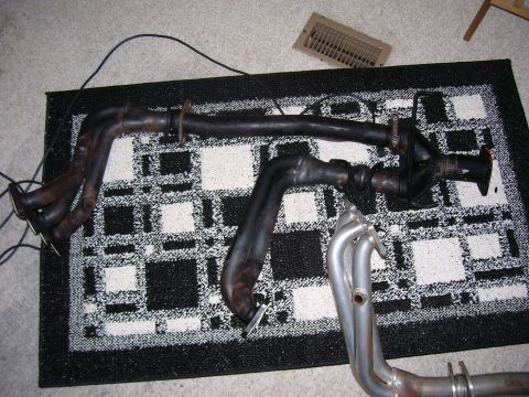 1993 Mazda MX-6 - Two sets of exhaust headers are laid out on a patterned rug, one black and one silver, with a Supermotors.net logo visible.