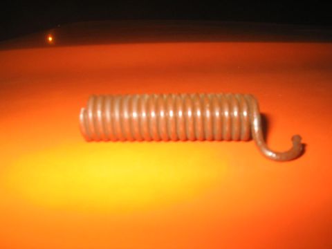 1979 Ford Bronco - Close-up of a small zinc-plated coil extension spring with a hooked end, resting on an orange painted body panel. The shot is slightly blur…