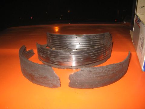 1979 Ford Bronco - Night shot on an orange painted surface showing a worn drum brake assembly: two slotted and drilled steel shoe tables with the friction lin…