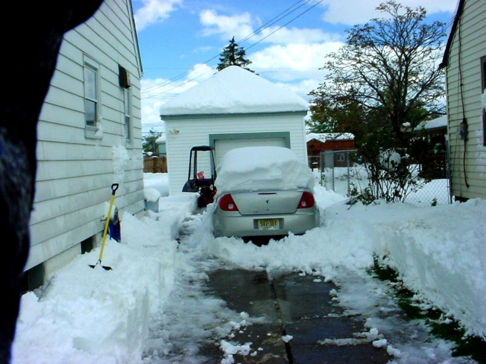 Silver sedan with snow piled high on its roof and hood, parked in a driveway next to a snow-covered garage