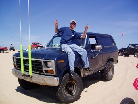 1986 Ford Bronco - A man poses on the hood of a dark blue Ford Bronco on a sunny day at the beach.