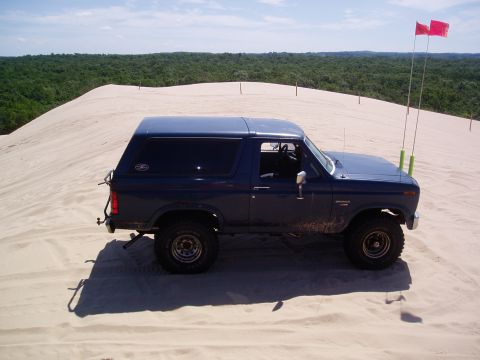 1986 Ford Bronco - A blue Ford Bronco is parked on a sand dune under a partly cloudy sky.