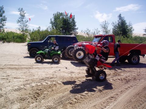 1986 Ford Bronco - Two ATVs are being ridden on sand dunes near a dark blue Ford Bronco and a red pickup truck.