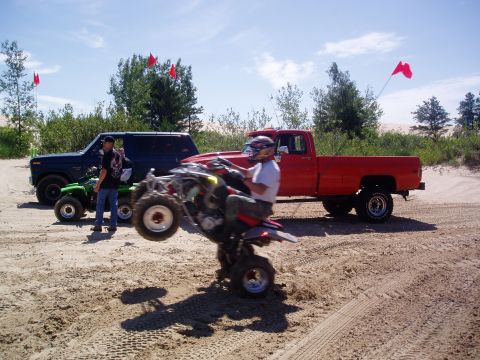 1986 Ford Bronco - A red pickup truck and a blue SUV are parked on a sandy dune while ATVs are being ridden.