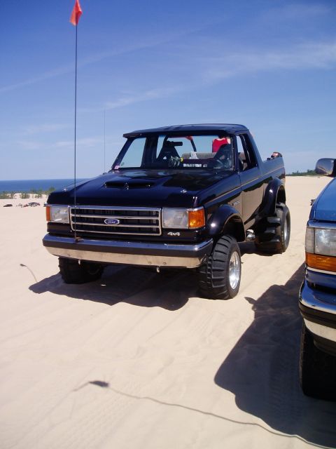 1986 Ford Bronco - A black off-road SUV with large tires is parked on a sandy dune under a clear blue sky.