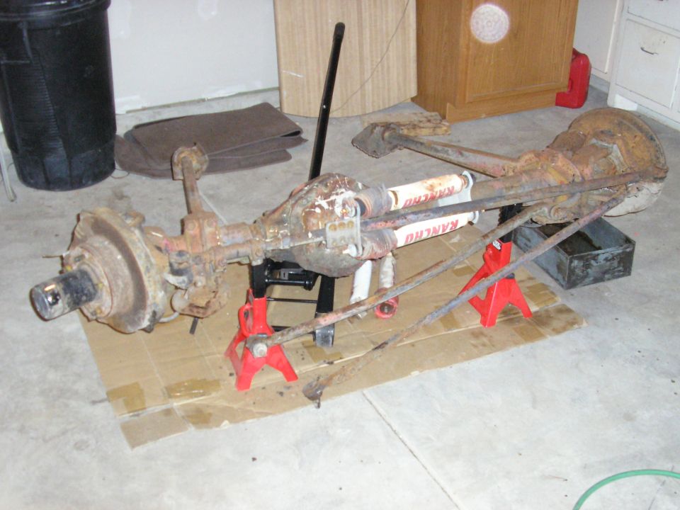 Removed rusty solid front axle with white Rancho shocks on red jack stands in a garage