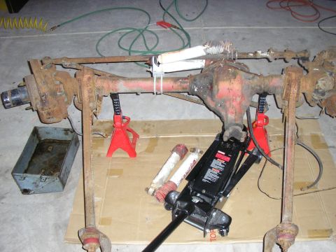 1996 Ford Bronco - A rusty front axle assembly is supported by jack stands and a floor jack on a cardboard-covered floor.