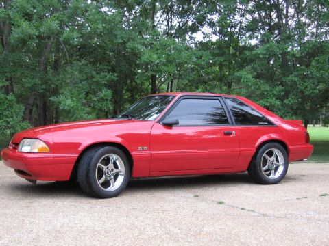 1967 Ford Mustang - A red coupe with a 5.0 badge is parked outside on a gravel surface with trees in the background.