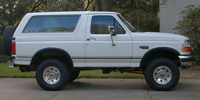 White Ford Bronco SUV in side view, lifted with chrome wheels and rear-mounted spare tire.