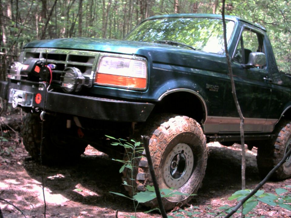 Green lifted Ford Bronco with winch bumper and mud tires in woods, front-left view
