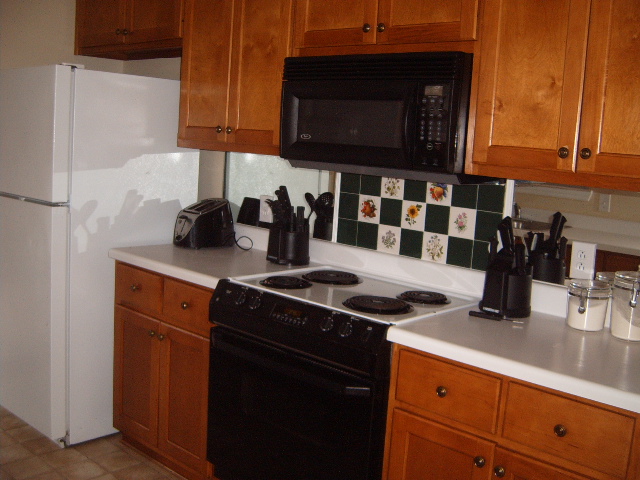 Kitchen with white refrigerator, black stove, black microwave, and wood cabinets. Tile backsplash behind stove