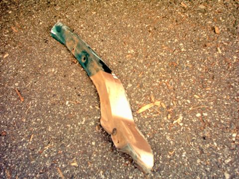 1996 Ford Bronco - A detached fender trim piece, showing green and tan paint, lies on a gravelly asphalt surface.