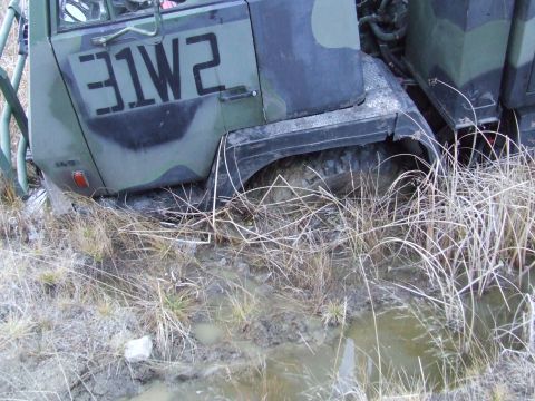 1990 Ford Bronco - A military truck with camouflage paint is stuck in muddy, grassy terrain, with its front tire partially submerged.