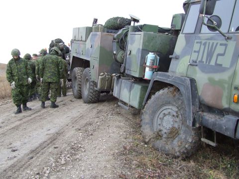 1990 Ford Bronco - Several soldiers stand near a large, camouflaged military truck on a dirt road.