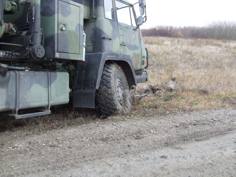 1990 Ford Bronco - A camouflaged military truck is partially stuck in the mud on a dirt road, with dry grass and trees in the background.