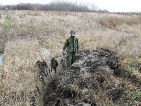 1990 Ford Bronco - A soldier in camouflage stands in a muddy ditch, surrounded by dry grass and vegetation under an overcast sky.