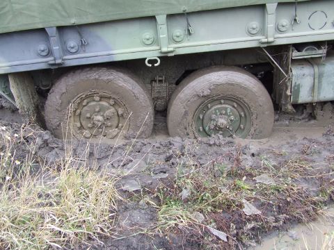 1990 Ford Bronco - A military truck is stuck in thick mud, with its wheels submerged and surrounded by wet grass and debris.
