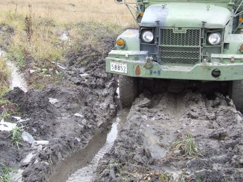 1990 Ford Bronco - A military truck is stuck in a muddy rut, with its front tires deeply embedded in the mire.
