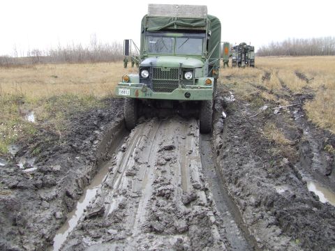 1990 Ford Bronco - A military truck navigates a challenging muddy terrain, with a second vehicle visible in the background.