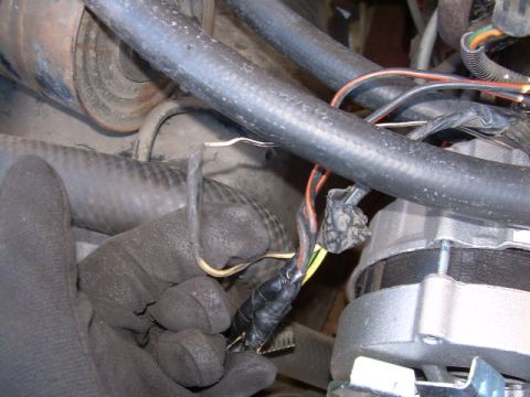1988 Ford Bronco - gloved hand holds a taped wiring harness connector with colored wires near an alternator and rubber hoses inside an engine compartment.