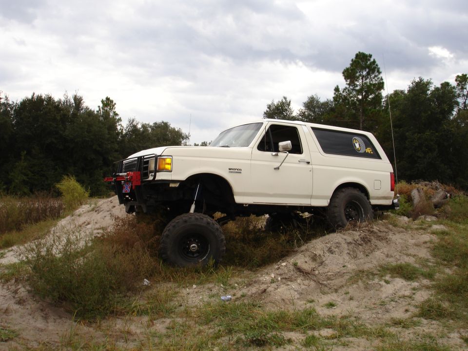 White 1989 Ford Bronco with lifted suspension and off-road tires driving on a dirt hill