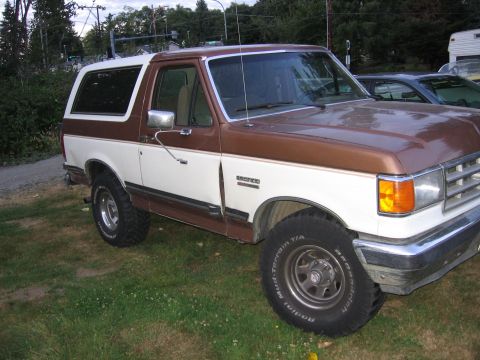 1987 Ford Bronco - A two-tone brown and white 1987 Ford Bronco is shown from the front passenger side, parked outdoors on a grassy area.