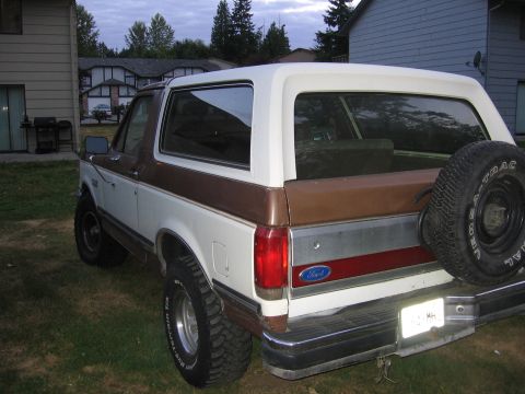 1987 Ford Bronco - The rear of a two-tone Ford Bronco is parked on a grassy lawn in front of residential buildings.