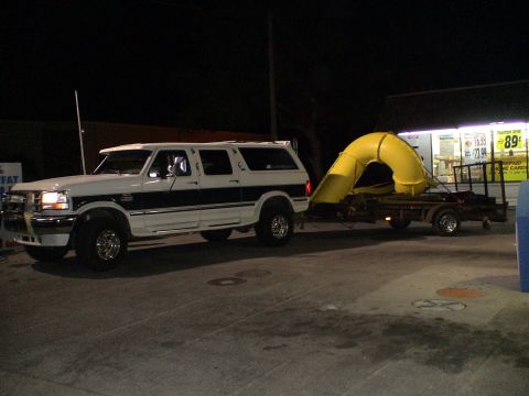 1993 Ford C-350 (Centurion Conversions) - This white and dark green SUV tows a large yellow slide on a trailer at night, possibly after an event.
