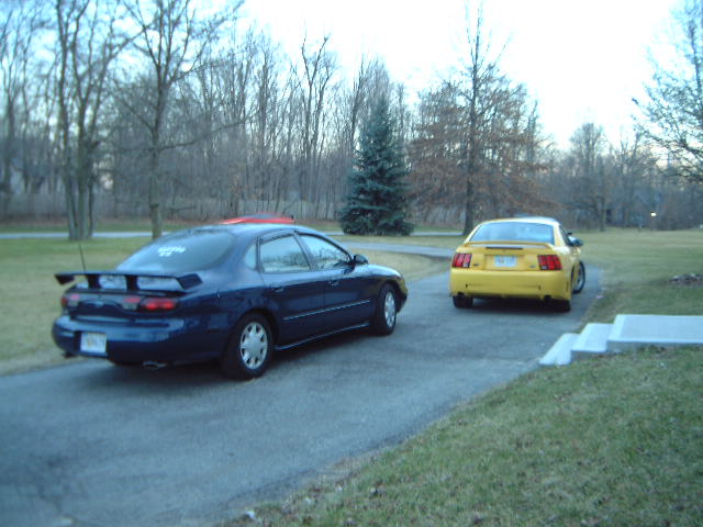 Dark blue Ford Taurus sedan with rear spoiler parked behind a yellow Mustang in a driveway