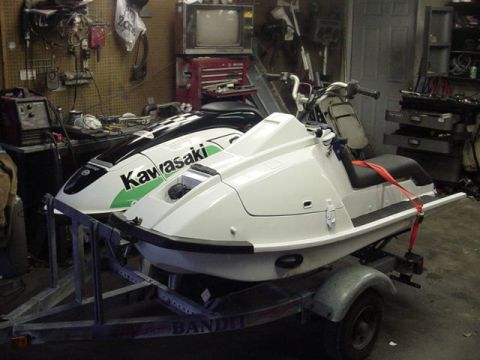 1995 Ford Mustang - Two jet skis are parked on a trailer inside a cluttered garage. One jet ski is white and the other has green and black Kawasaki graphics.