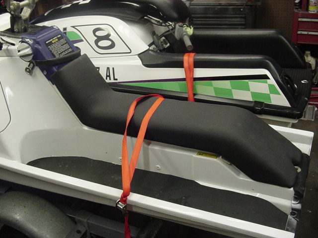 Two jet skis are strapped down on a trailer. One has a black seat and orange straps. The other has a white body with green checkered pattern and the number 8
