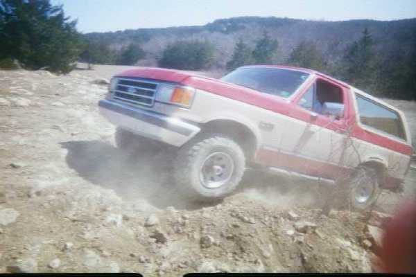 Red and white 1987 Ford Bronco driving off-road, kicking up dust