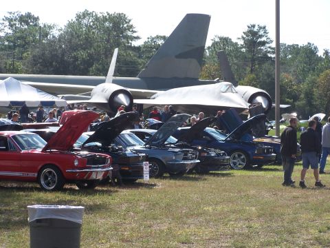 1973 Ford Ranchero - A row of classic cars, including a red 1965 Ford Mustang GT, are displayed with their hoods open at an outdoor event, with a large aircraft…