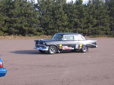 1989 Ford Mustang - A black 1956 Chevrolet sedan, adorned with racing decals, sits on an asphalt surface with trees in the background.